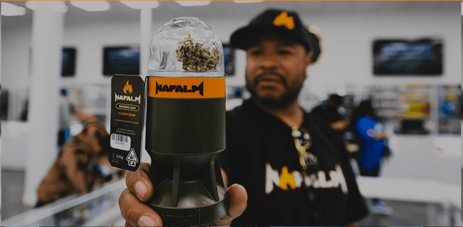 Man holding a grenade-shaped Napalm cannabis flower bomb container in a blurred dispensary setting.